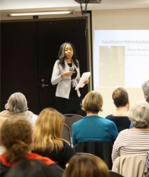 A woman speaks in front of a crowd with a presentation in the background