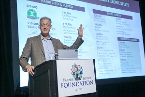 A man at a podium gestures to some figures on a screen behind him