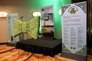 A flower arrangement in the shape of a bowtie is set up in front of a stage with several large signs